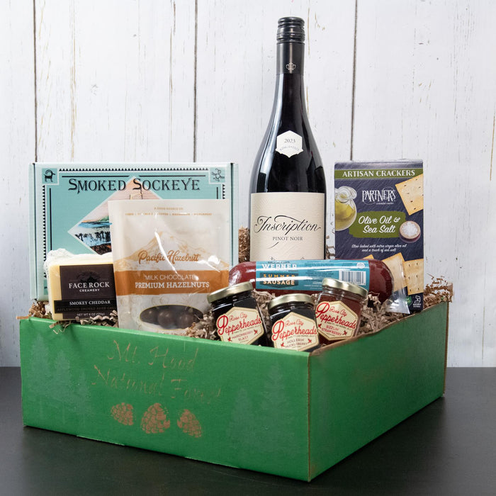 A gift basket containing a bottle of King Estate Pinot Noir, smoked sockeye salmon, summer sausage, cheddar cheese, hazelnuts covered in milk chocolate, and pepper jelly jars, all packaged in a green eco-friendly gift box.