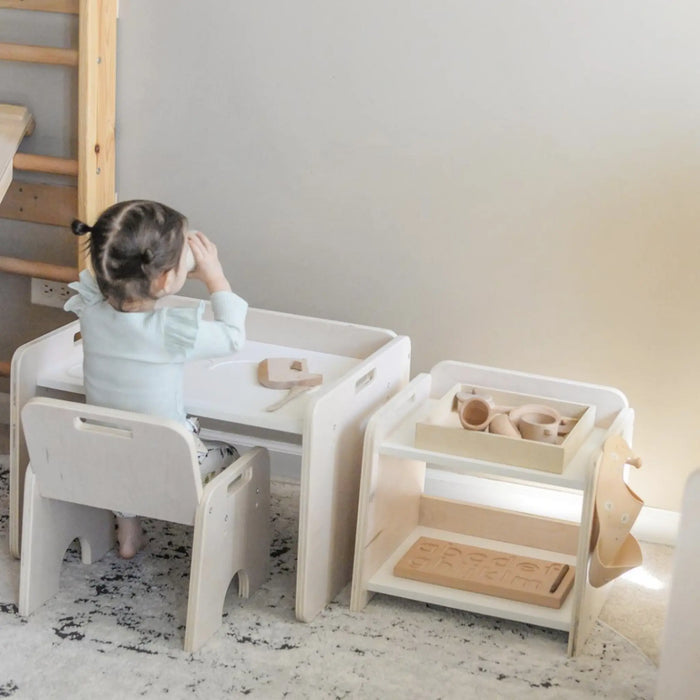 Toddler girl sitting on a wooden montessori chair from Sapiens Child in front of wooden montessori weaning table drinking from cup. Beside the table is the Papaya shelf by Sapiens Child . The shelf has a wooden tray with wooden toys and a hook on the side that holds a beige bib.