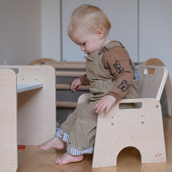 Side view of a blonde toddler boy in corduroy overalls sitting barefoot on an adjustable Sapiens Child wooden Montessori weaning chair next to a matching weaning table on a hardwood floor.