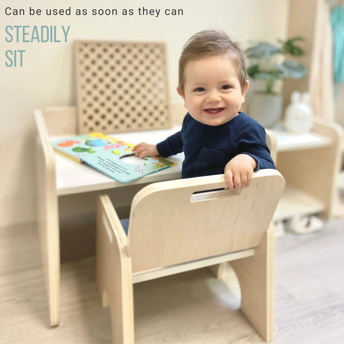 little one year old smiling boy with blue shirt sitting on the weaning chair from Sapiens Child. View is from behing but he is turning around and smiling. He was reading a book on top of the montessori activity table. The piece looks beautiful and steady.