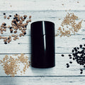 Small black seed storage vault container surrounded by various seeds on white wooden surface